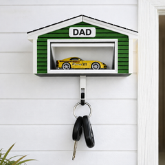 A green house-shaped wall-mounted key holder with a white trim and the word 'DAD' on the front, displaying a yellow race car inside. A metal key hook hangs below, holding two keys, against a white paneled wall.